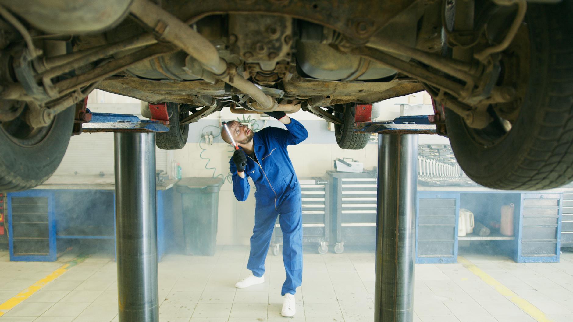 Mechanik w niebieskim kombinezonie ogląda od spodu samochód na podnośniku