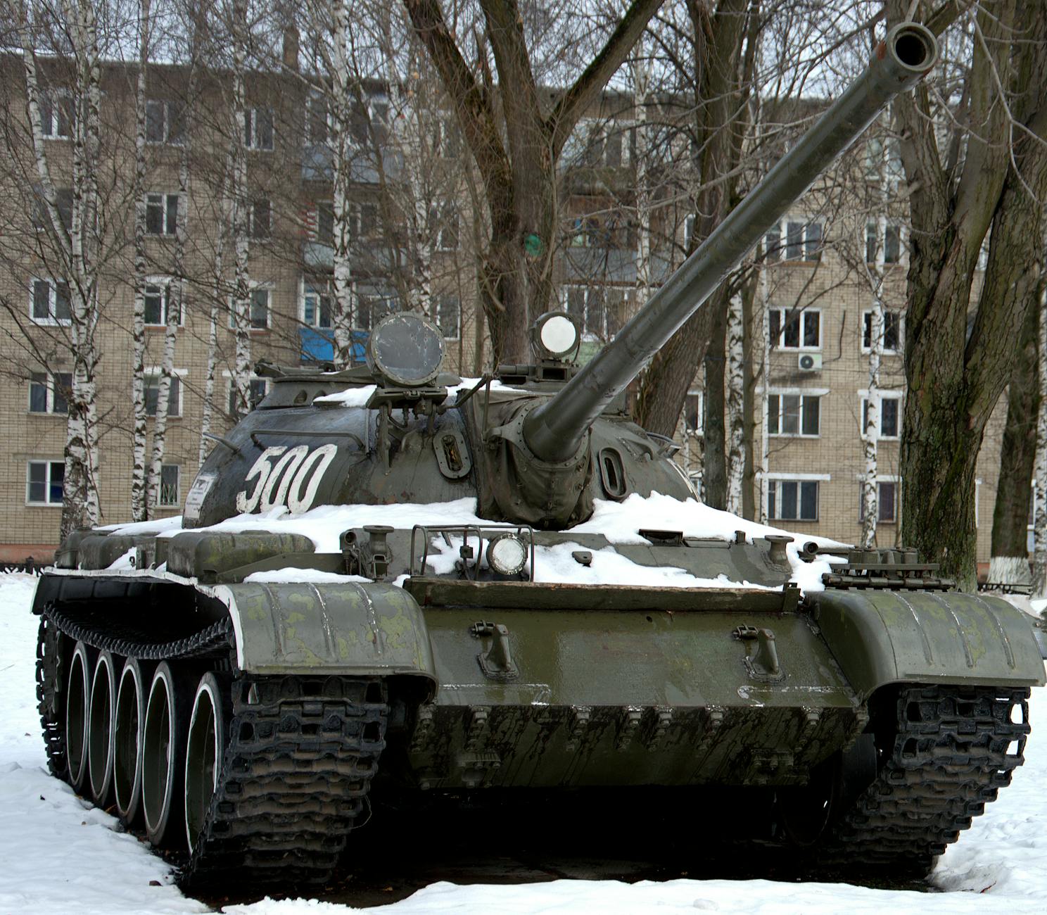 Czołg T-72M1 od przodu na ośnieżonym placu między drzewami i budynkami