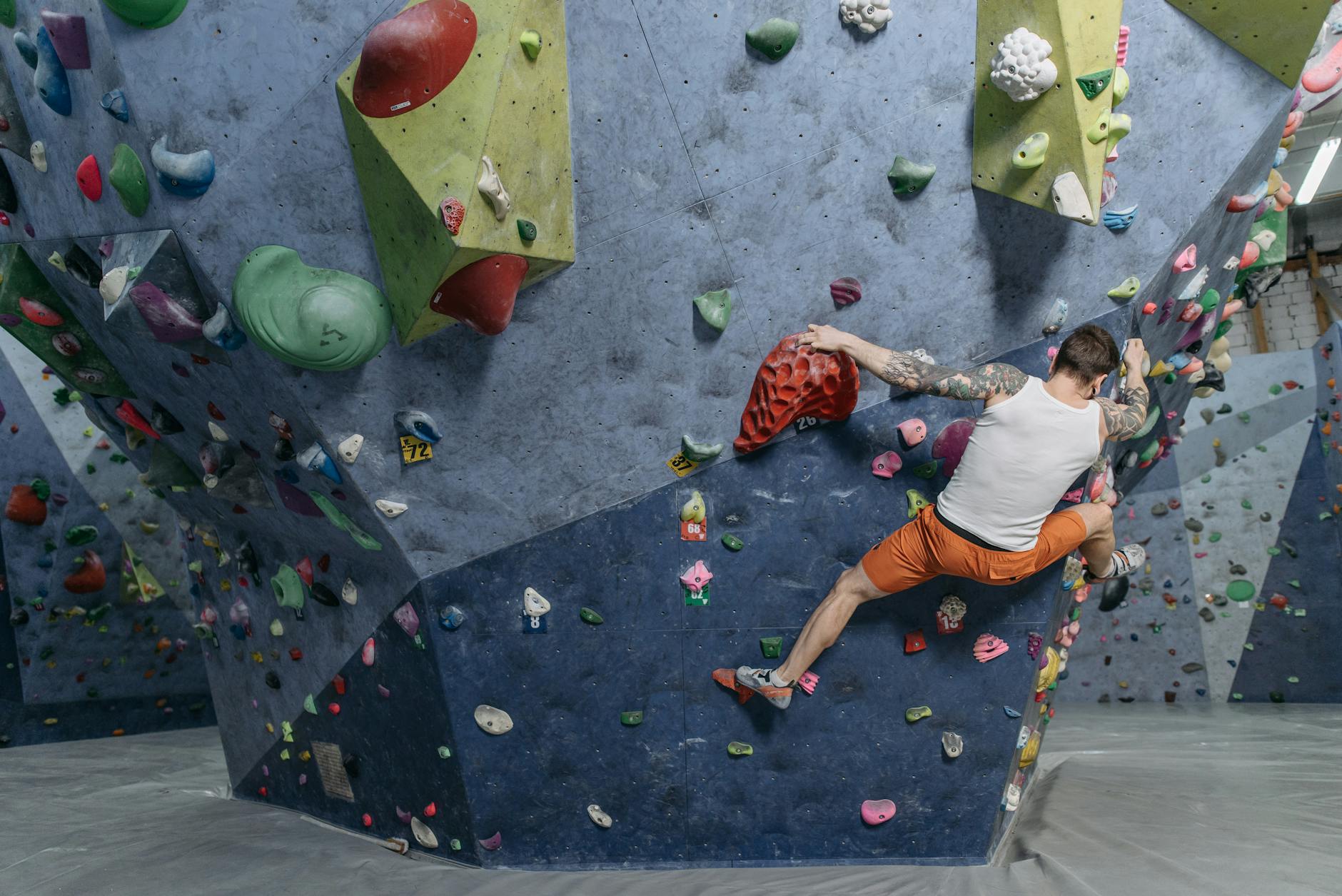 Wytatuowany mężczyzna wspina się na kolornej ściance boulderowej w hali